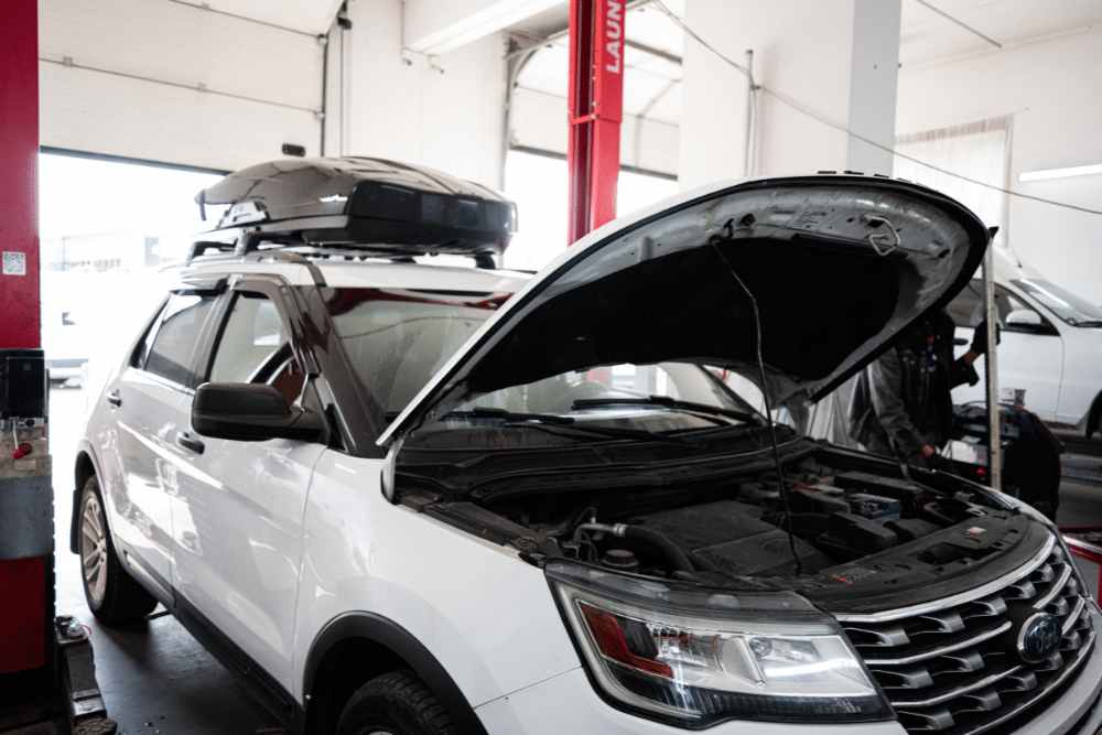 Ford repair and service in Floyds Knobs, IN at Schindler's Garage. A white Ford SUV with its hood open inside an auto repair shop, parked near a red vehicle lift. A roof cargo box is mounted on top of the vehicle.