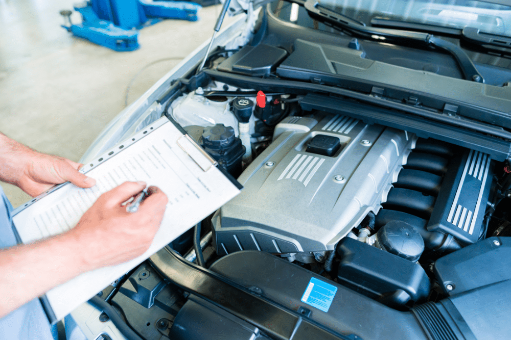 Car Maintenance Checklist in Floyds Knobs, IN at Schindler's Garage. Mechanic inspecting a car engine with the hood open and filling out a checklist on a clipboard in an auto repair shop.