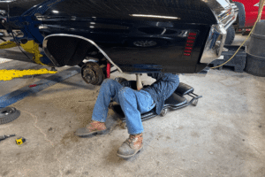 Spring car maintenance, auto repair in Floyds Knobs, IN by Schindler’s Garage. Image of a technician working beneath a lifted classic muscle car in a professional auto repair shop, highlighting skilled maintenance, restoration support, and dependable automotive care.