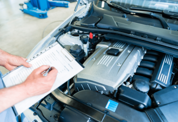 Car Maintenance Checklist in Floyds Knobs, IN at Schindler's Garage. Mechanic inspecting a car engine with the hood open and filling out a checklist on a clipboard in an auto repair shop.